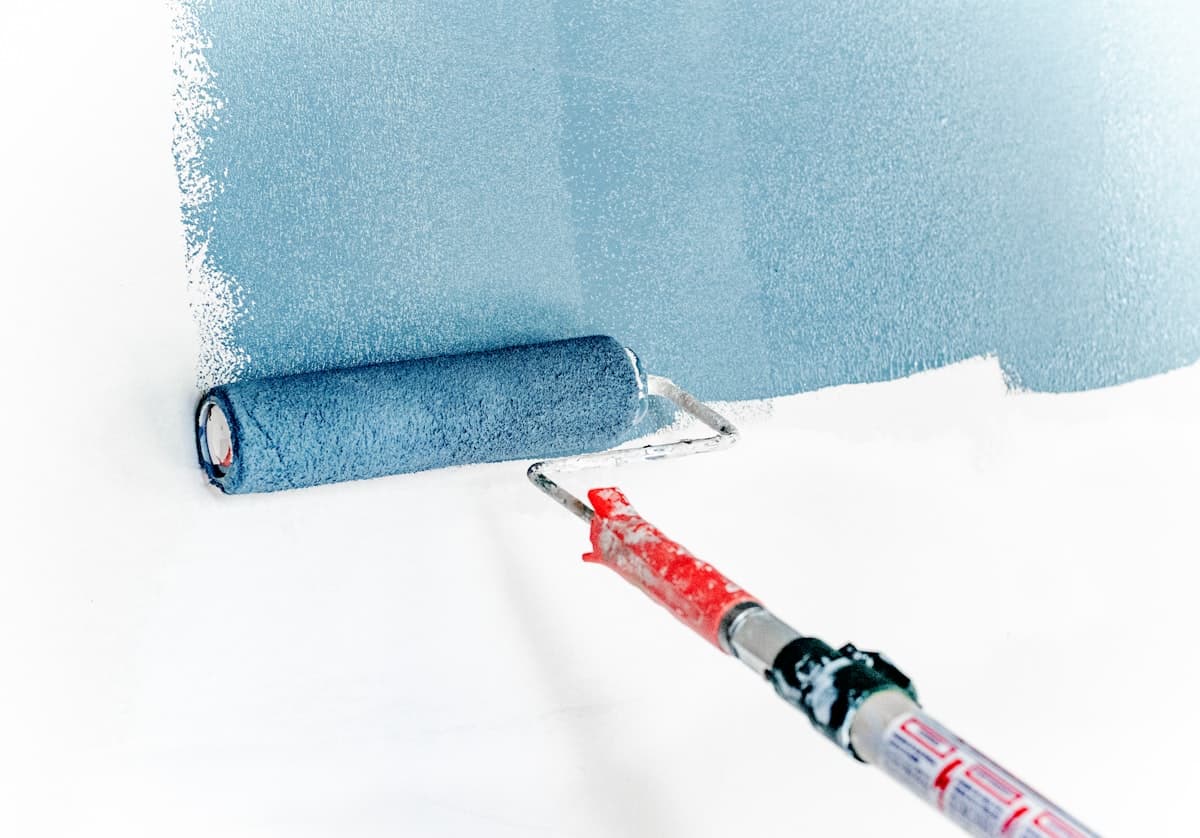 A paint roller on an extension pole part way through a first coat, blue-grey going on over white. Illustration, not a Construction Hub job.