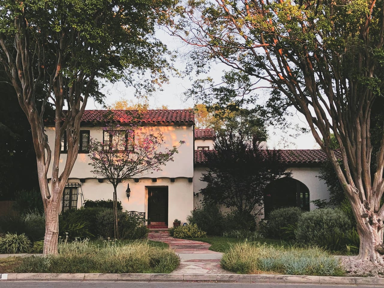 A two-story white stucco house with a red tile roof behind two street trees
