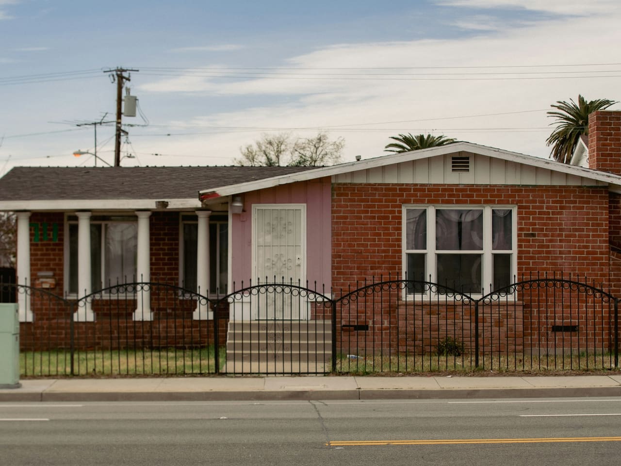 A single-story red brick house behind a low iron fence on a quiet residential street