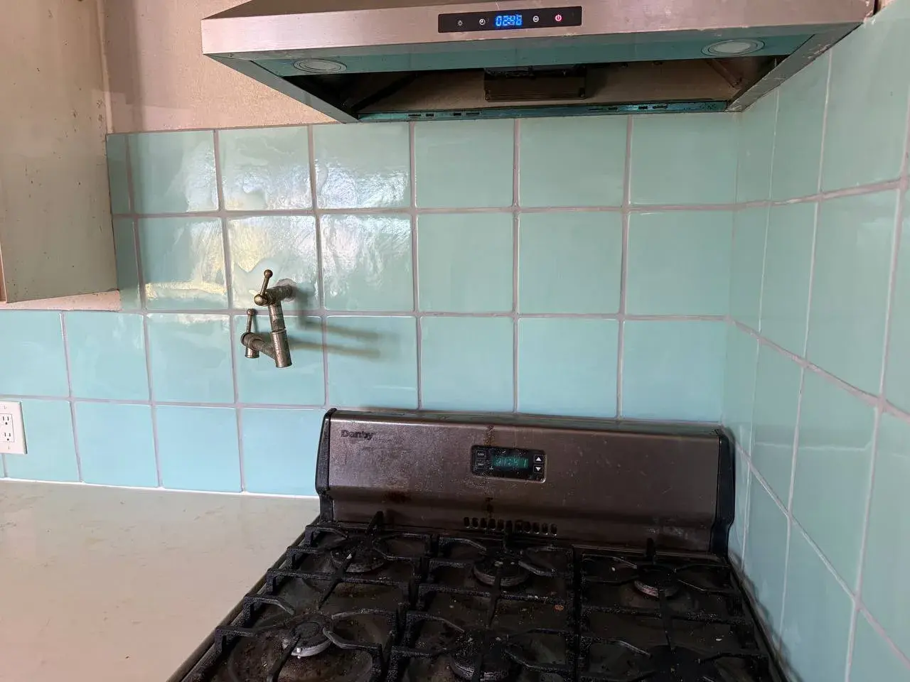 Teal square tile behind a gas range, around a brass pot filler and up to the stainless hood. After the work, by Construction Hub in Los Angeles.