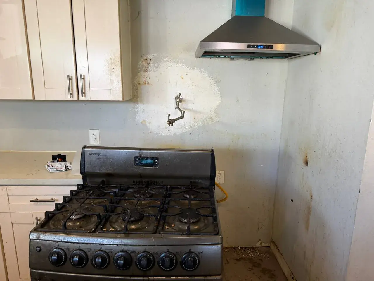 Bare stained wall behind a gas range, patched around a brass pot filler, stainless hood above. Before the work, by Construction Hub in Los Angeles.