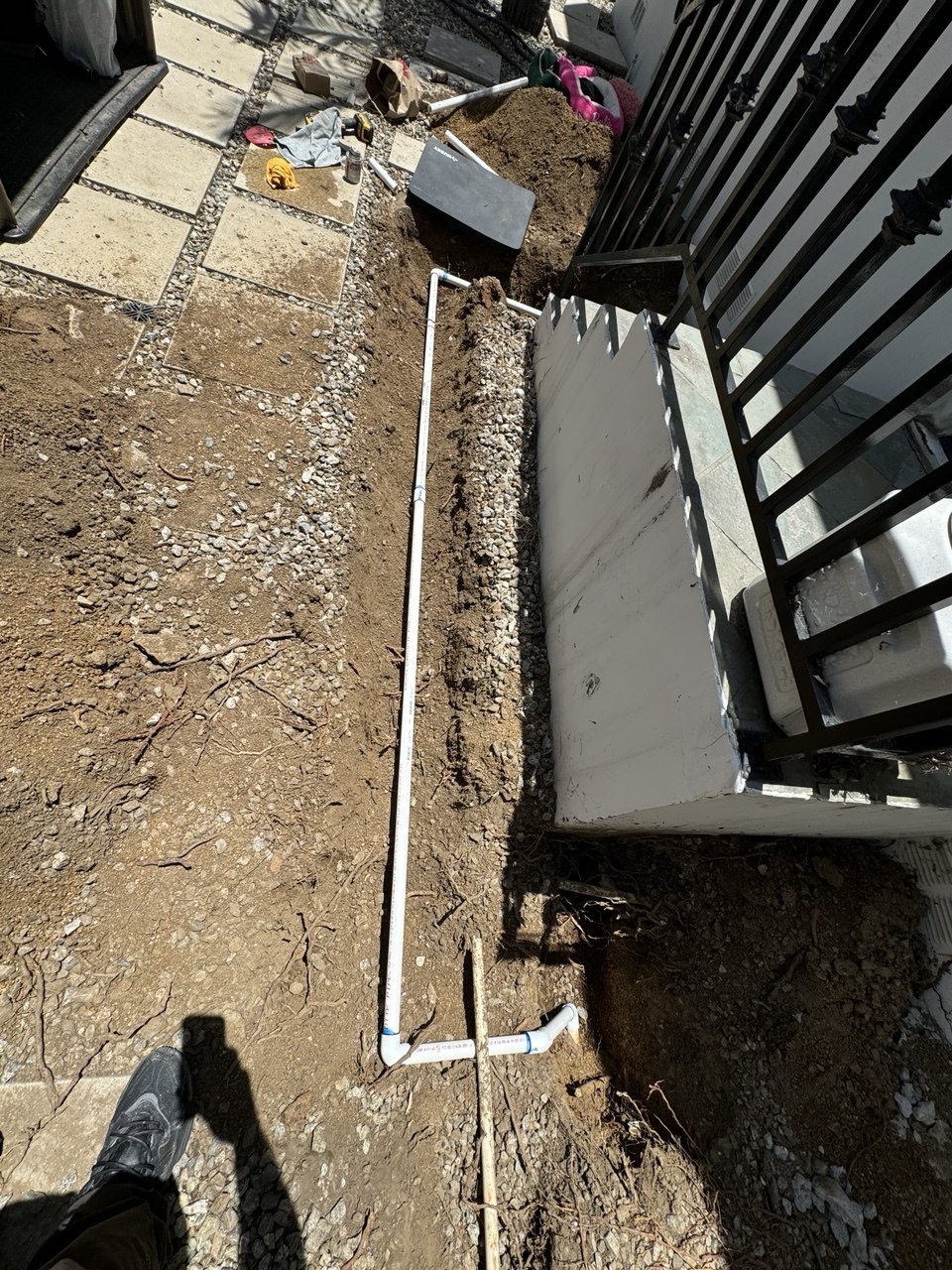 A new PVC line laid in an open trench beside a white garden wall and an iron railing, an elbow turned up at the near end. Fine roots still show in the dirt on the left.