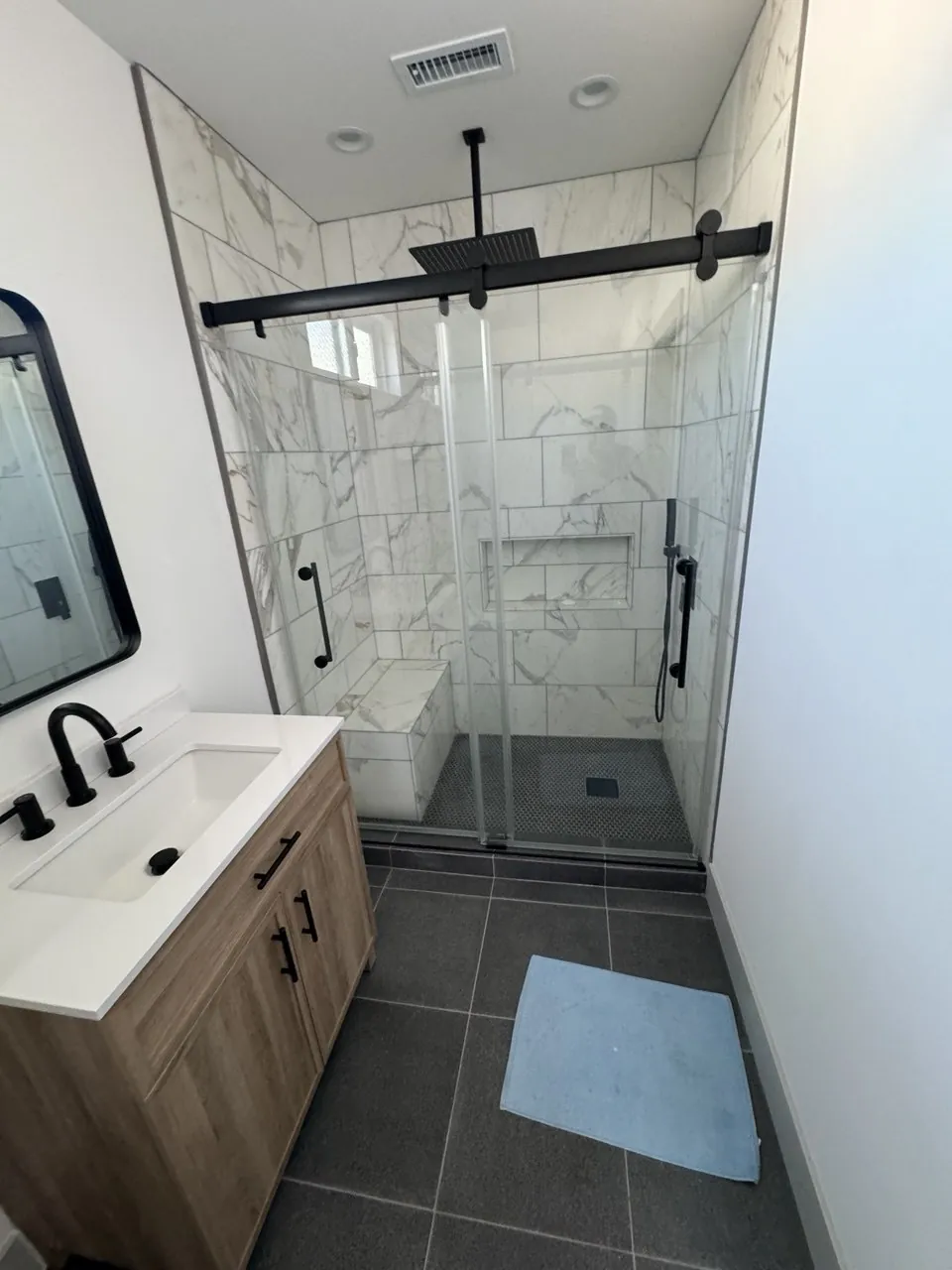 Remodeled bathroom with marble-look tile shower, black sliding glass door and wood vanity