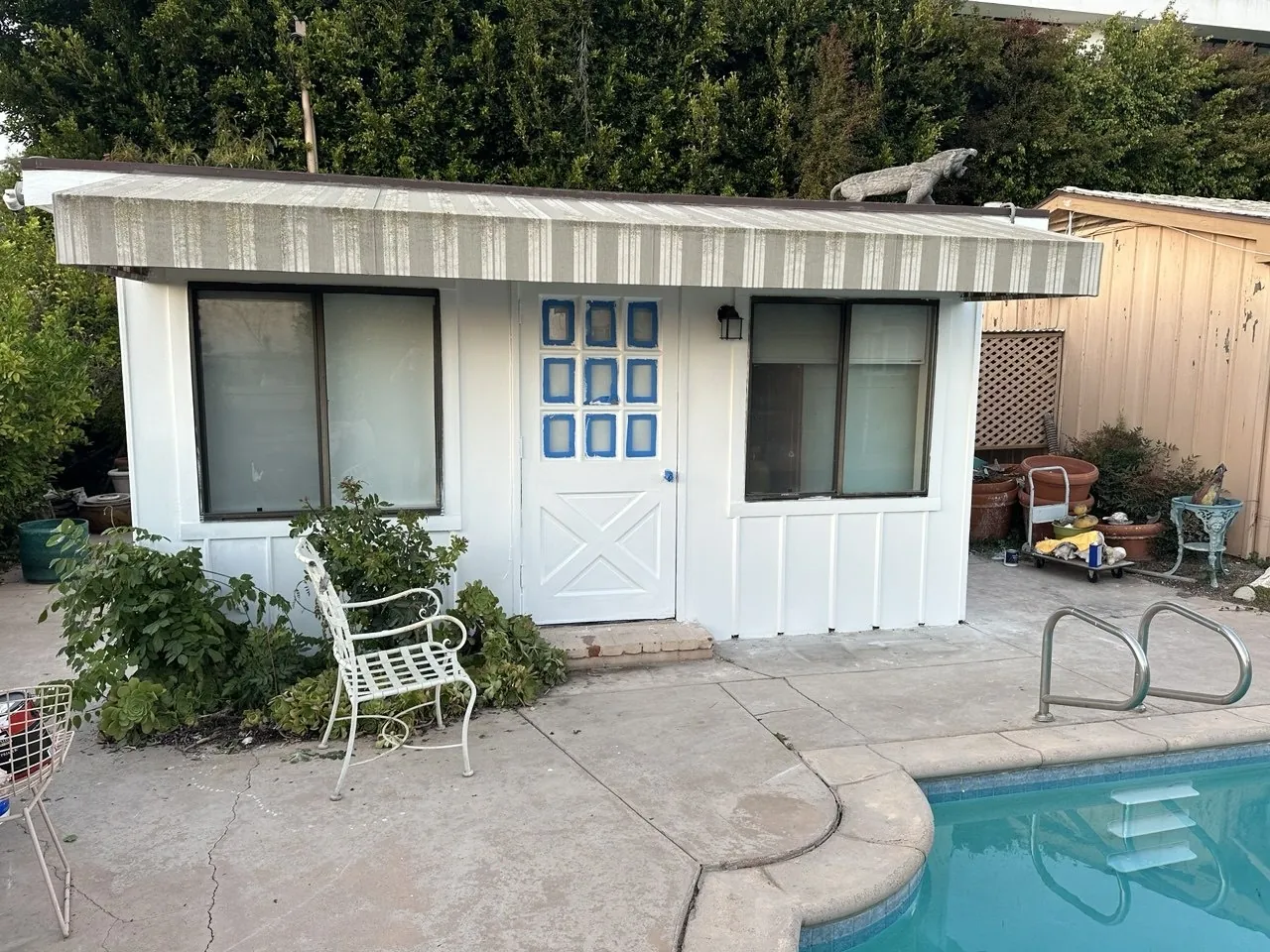 Freshly painted white pool house with board-and-batten siding and blue window accents on the door